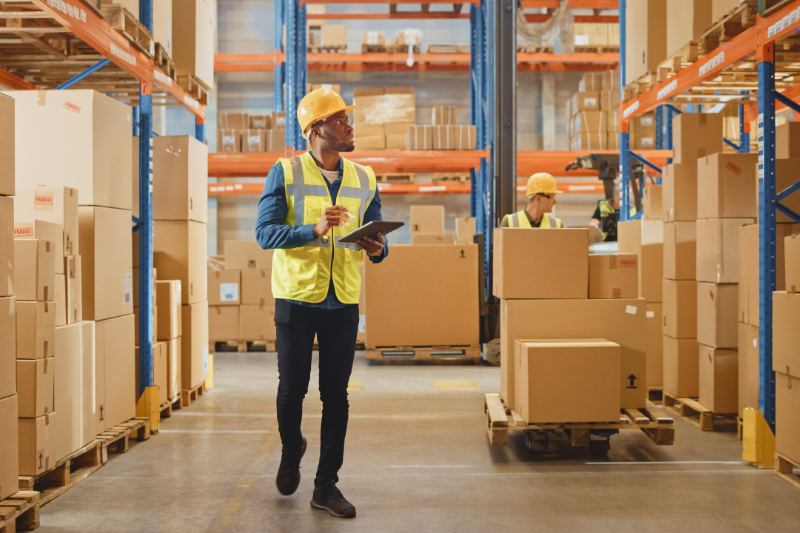 Handsome Male Worker Wearing Hard Hat Holding Digital Tablet Computer Walking Through Retail Warehouse full of Shelves with Goods. Working in Logistics and Distribution Center.